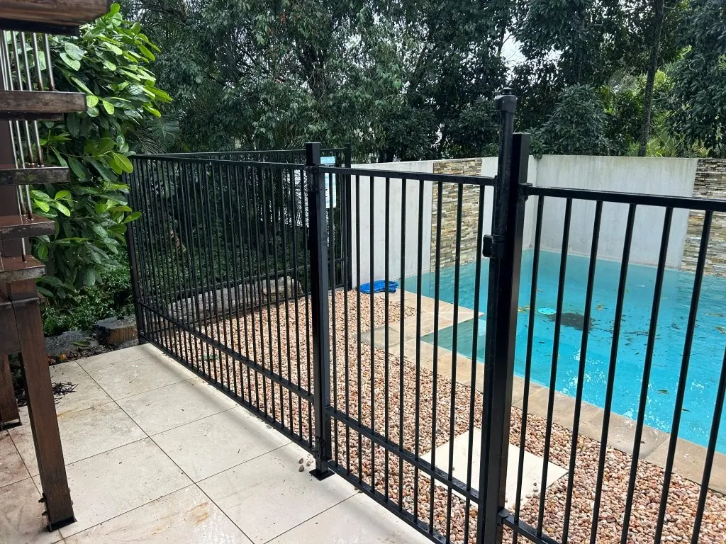 Black aluminium pool fence and self-closing gate around a pool with pebble surrounds
