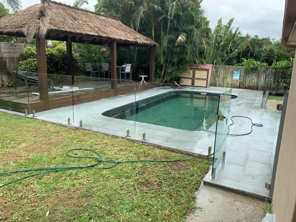 Frameless glass pool fence with gold spigots around a pool and thatched gazebo